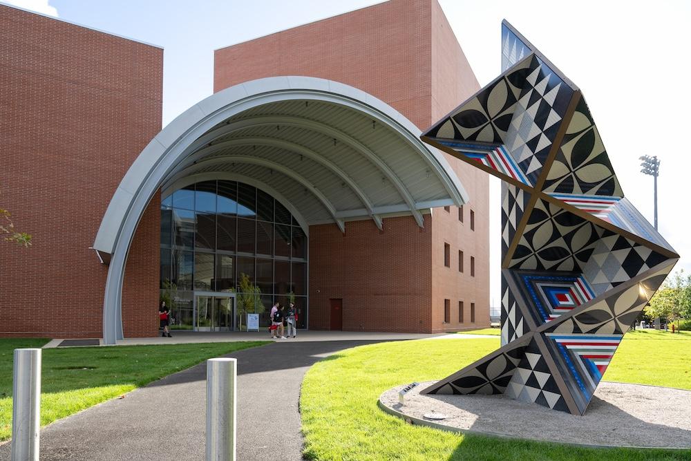 The Edward and Joyce Linde Music Building - a modern brick building with a large, curved metal entrance is seen. In the foreground, there is a geometric sculpture featuring colorful, abstract patterns.