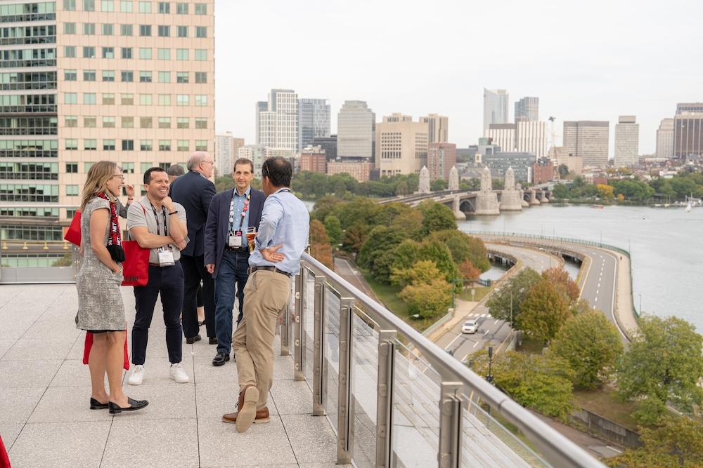 A group of people stands on a rooftop terrace overlooking the Charles River in Boston. The city skyline is visible in the background, with prominent buildings and a bridge crossing the river.