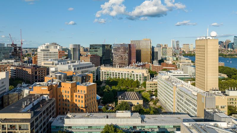 Aerial view of the MIT campus in Cambridge, featuring a mix of modern and traditional buildings. The Charles River and the Boston skyline are visible in the background under a partly cloudy sky.