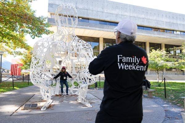 A person is taking a photo of another individual posing inside the "Alchemist" sculpture at MIT. The photographer is wearing a shirt that reads "Family Weekend." The scene is set outdoors, with a modern building and trees in the background.