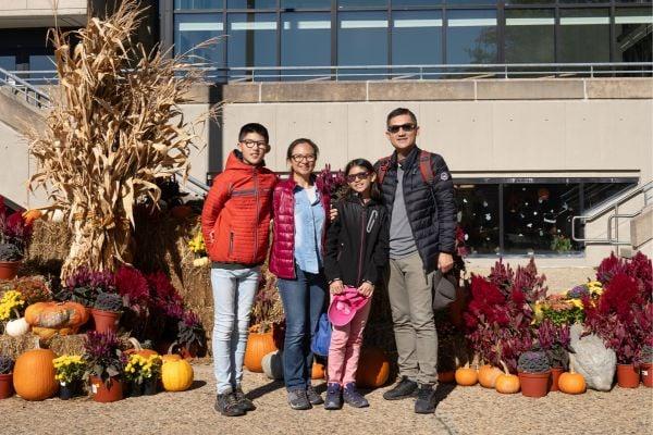 A group of four people standing together in front of a festive autumn display with pumpkins, cornstalks, and colorful plants. They are outdoors in a sunny setting, with a modern building and steps in the background.